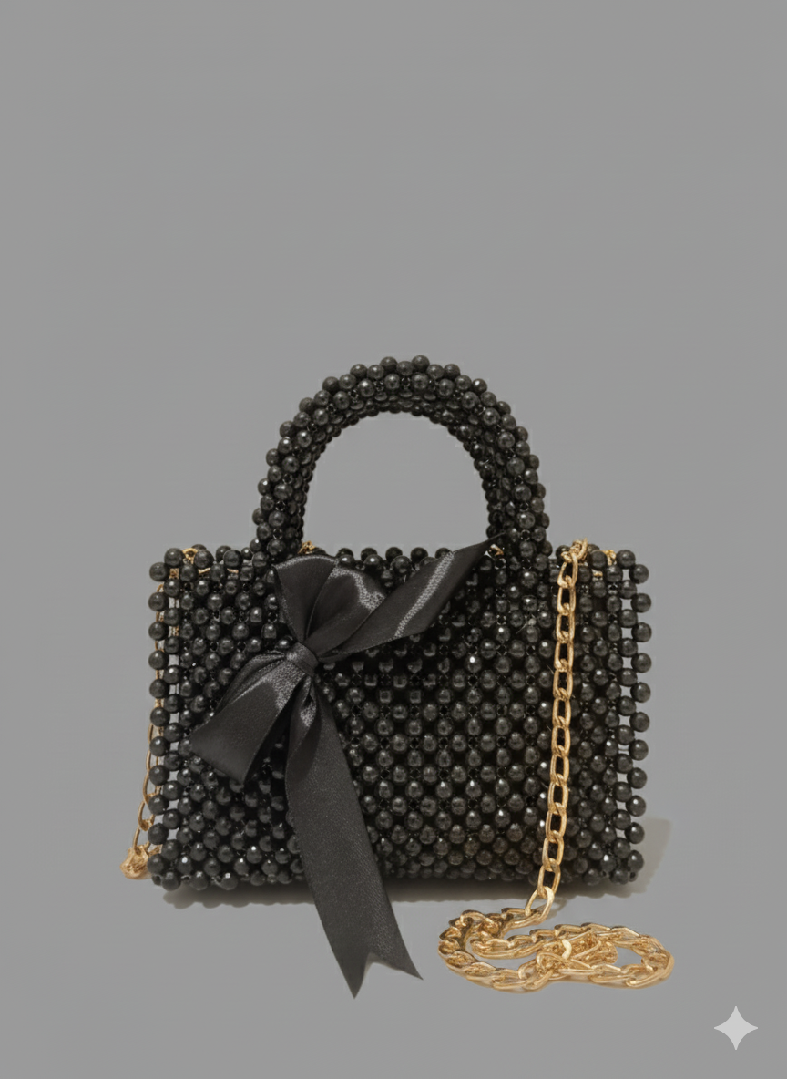  sophisticated black beaded handbag featuring a small top handle, a gold chain strap, and a prominent black satin ribbon tied at the front. The bag has a structured, glossy appearance.