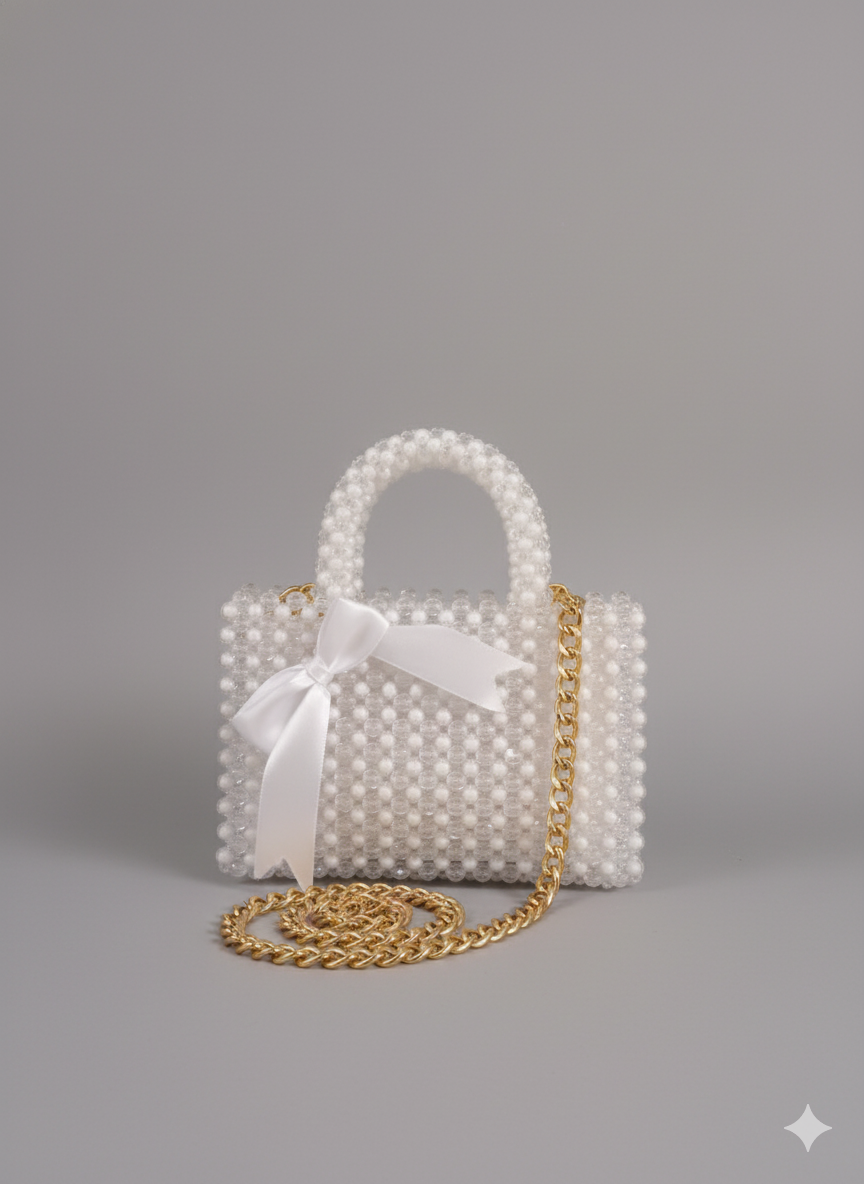 A small, white (clear) beaded handbag with a top handle, a gold chain strap, and a white satin ribbon elegantly tied onto the front.