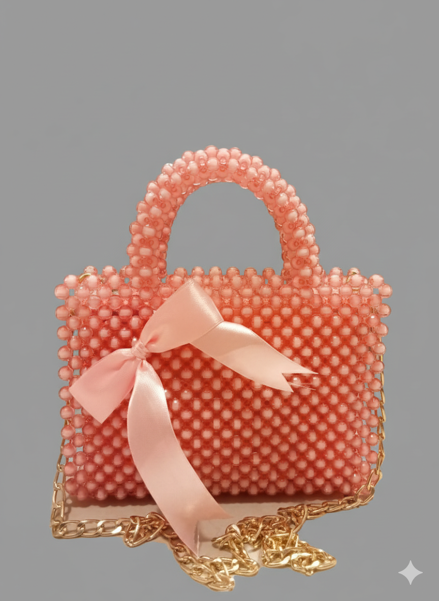 A small, peach-colored beaded handbag featuring a rounded top handle, a gold chain strap, and a loose, flowing pink satin ribbon tied at the front.